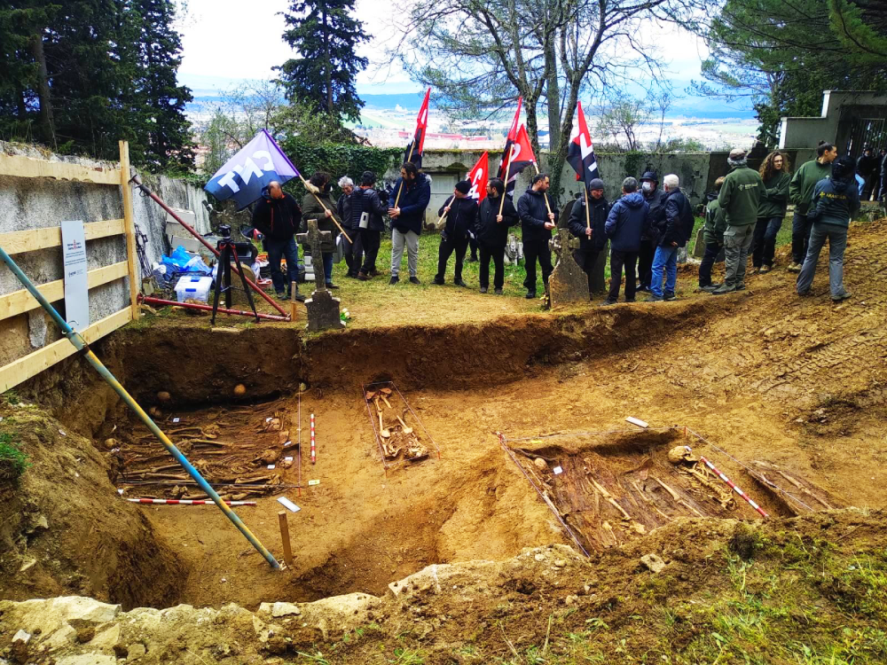 Foto de compañeras y compañeros de CNT enarbolando banderas en el cementerio de Berriozar. En primer plano, se observan los esqueletos de varios cuerpos exhumados; al fondo, trabajadoras de Aranzadi, familiares y otras personas.
