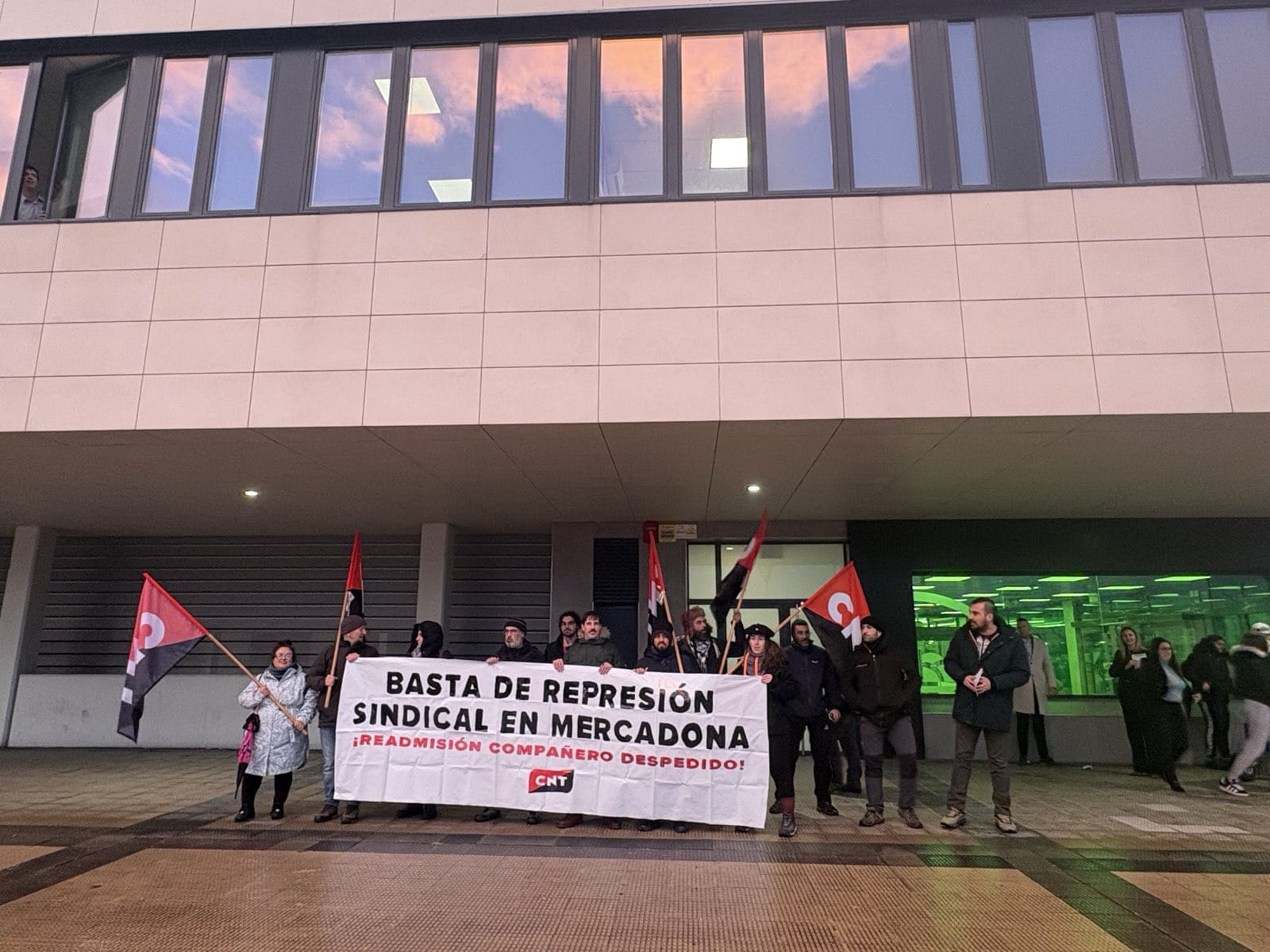 Militantes de CNT Iruñea portando banderas y una pancarta en la entrada a un Mercadona. En la pancarta se puede leer "Basta de represión sindical en Mercadona. Readmisión compañero despedido".