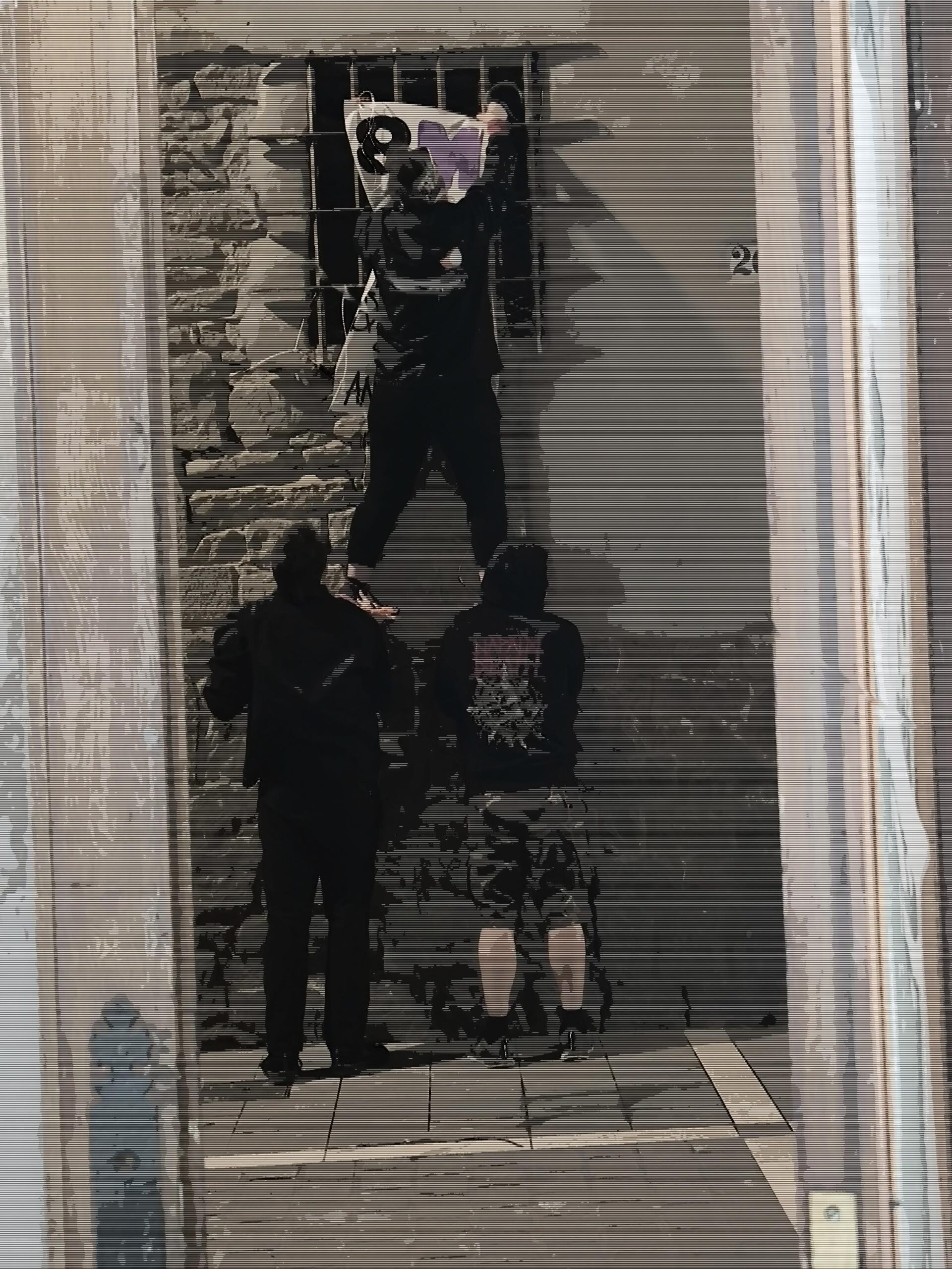 Una pared de piedra con una ventana enrejada. Tres personas con ropa oscura colaboran para colocar una pancarta rectangular en la parte superior de la ventana. Una de ellas se sostiene sobre otras dos para alcanzar la zona alta.