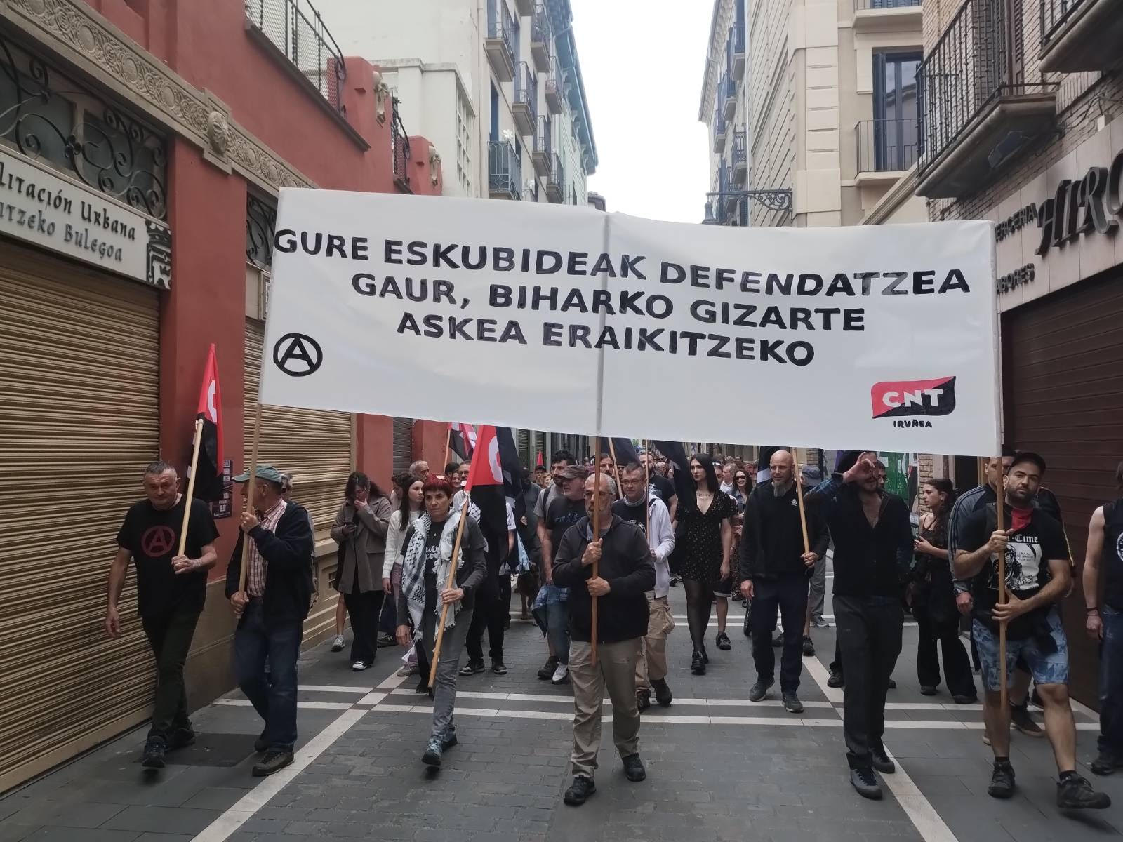 Manifestación del 1 de mayo organizada por CNT Iruñea. Un grupo de personas camina por una calle urbana portando una pancarta blanca con el lema en euskera 'Gure eskubideak defendatzea gaur, biharko gizarte askea eraikitzek' y el logotipo del sindicato.