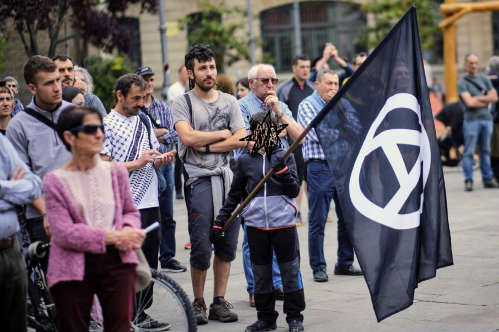 Personas de diferentes edades observan hacia su derecha con gesto pensativo y concentrado. Un niño sostiene una bandera negra con el símbolo de la A de anarquía.