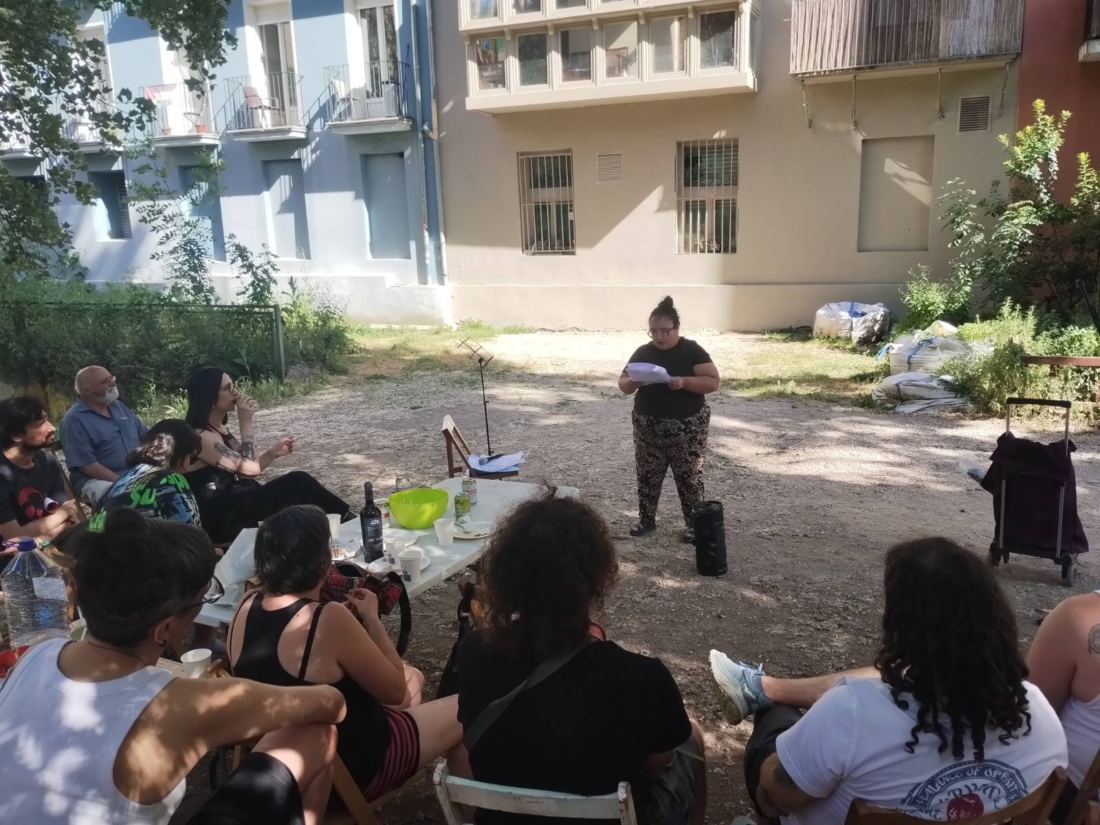 Una compañera de CNT Iruñea lee un relato bajo los árboles de una plaza. Ante ella, varias personas atienden a la lectura.